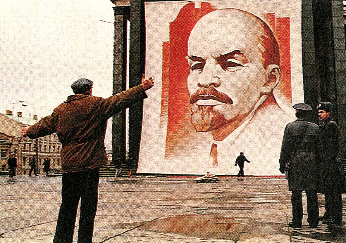 Large propaganda mural of Lenin on red background in Soviet Union, street scene with police and passerby during Cold War era, historical political art focus.