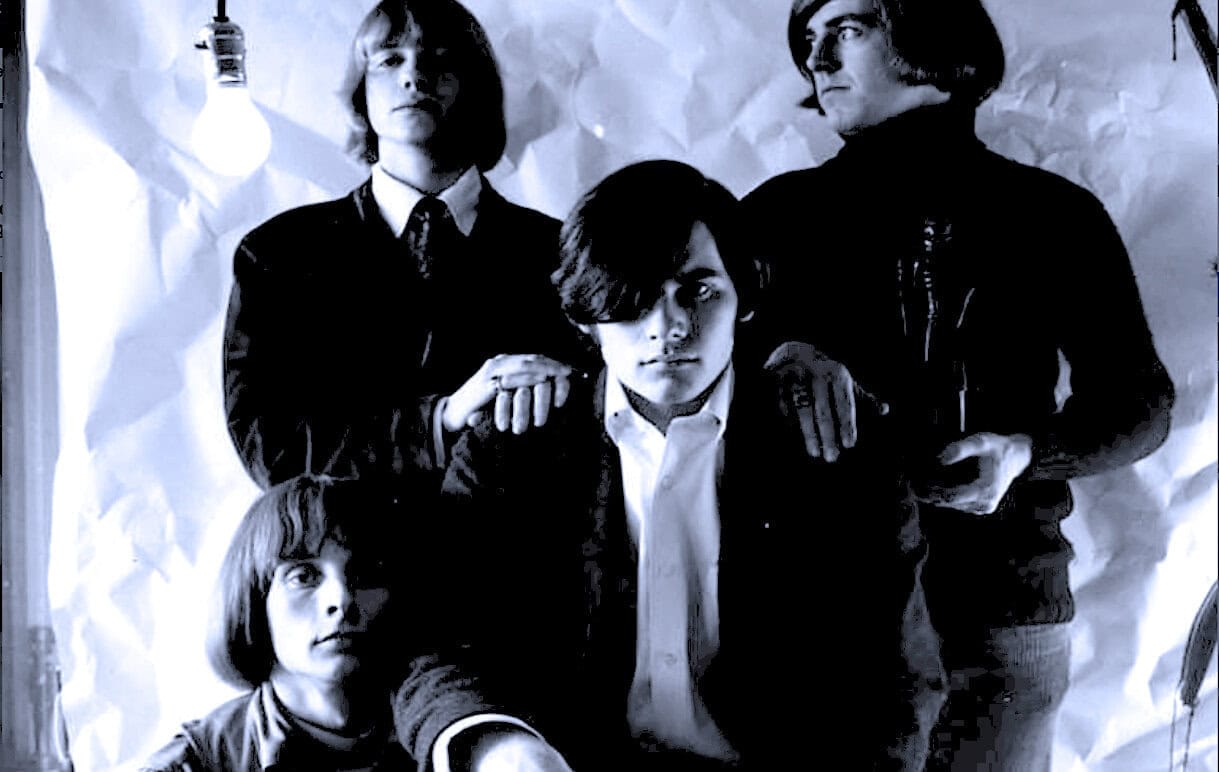 Black and white photo of a 1960s band with four members, stylish clothing, and intense expressions, representing music history.