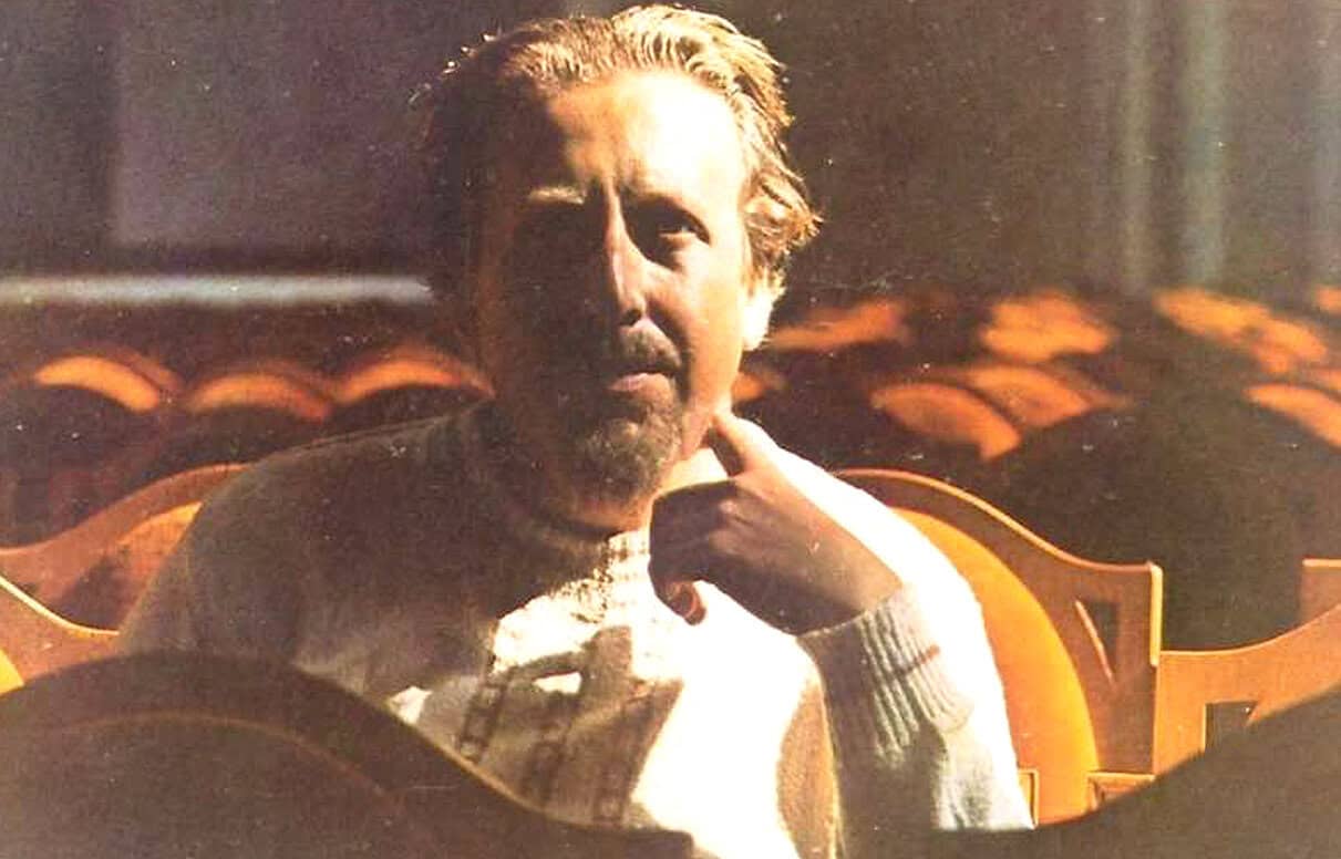 Thoughtful man sitting in an empty theater with rows of seats behind him, warm lighting creating a contemplative atmosphere, emphasizing reflection and nostalgia in a documentary or interview setting.