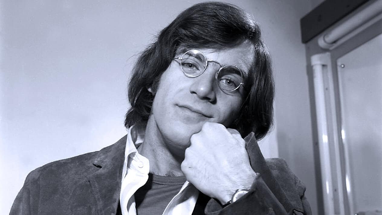 A black-and-white portrait of a man with shoulder-length hair, round glasses, and a thoughtful expression, representing music history and iconic voices featured in the Past Daily archive.