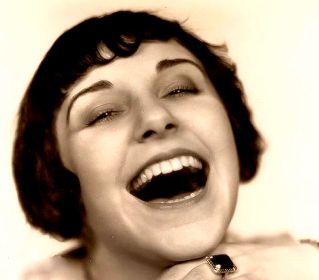Vintage black and white photograph of a woman with short, dark hair, laughing with her eyes closed, showcasing a joyful moment in a classic era, suitable for a historical music or news archive image.