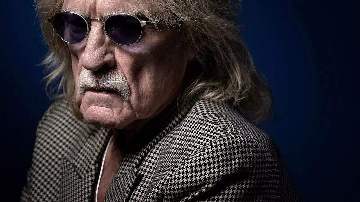 Close-up of an elderly man with long gray hair, wearing round sunglasses and a houndstooth jacket, expressing a serious or contemplative mood, suitable for historic news, music, and sound archive content.