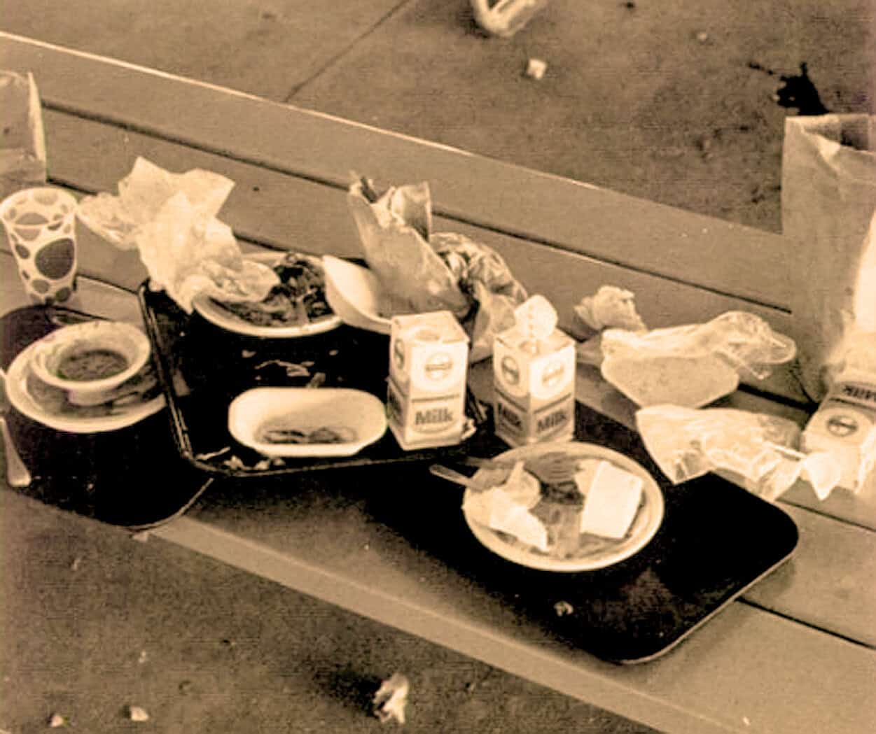 Discarded food and drink items on a picnic table, including milk cartons, paper plates with food, cups, and wrappers, in a messy outdoor setting for historical or nostalgic context.