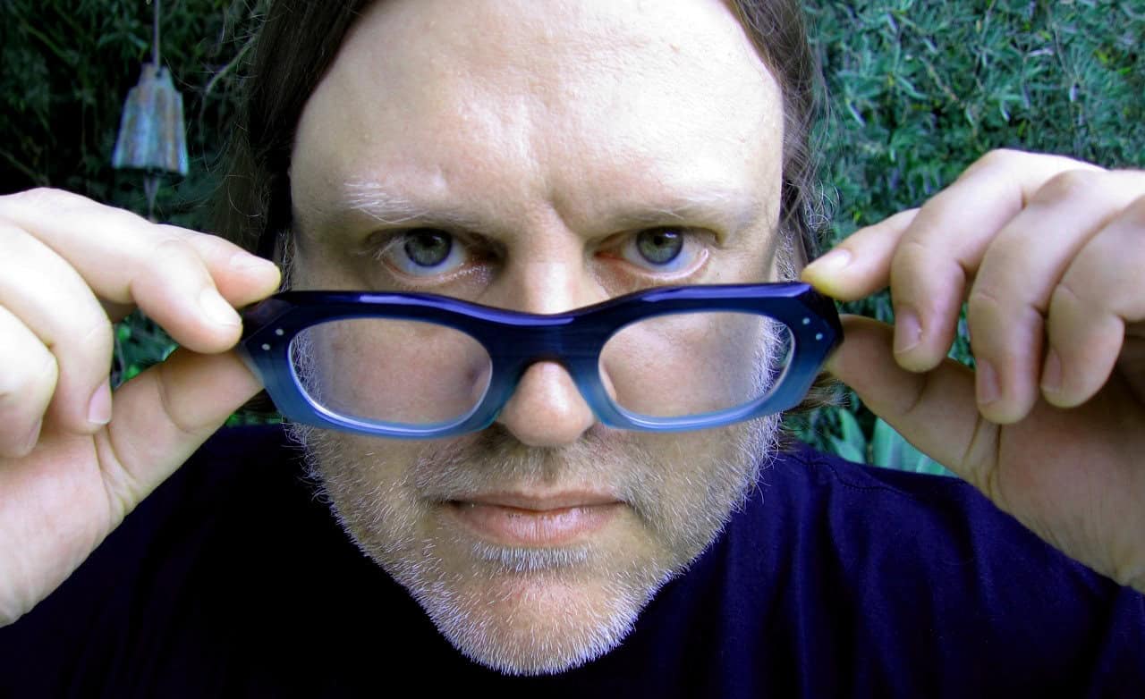 Close-up of a man with blue eyes holding sunglasses, outdoors in a garden, emphasizing focus and clarity in a natural setting.