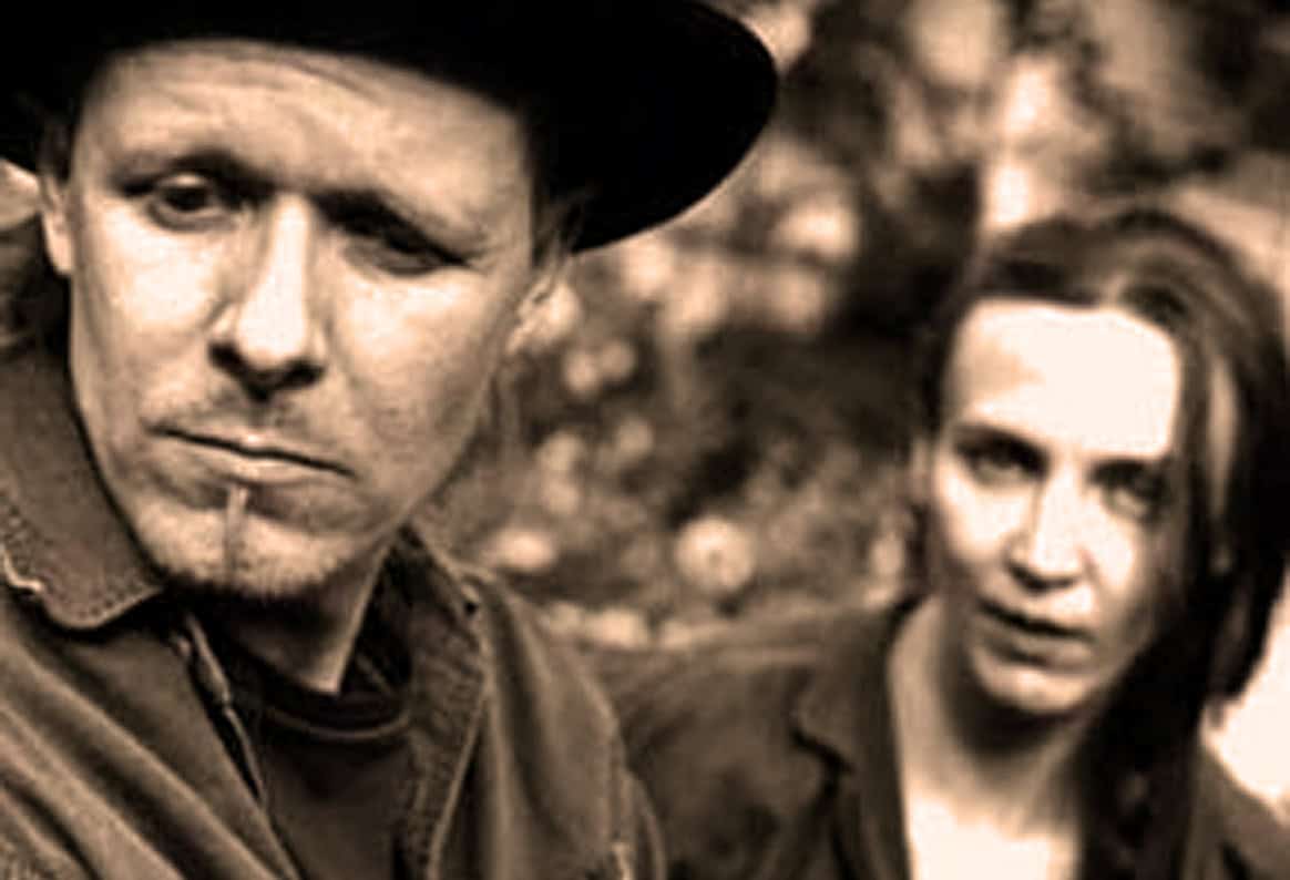 Sad man wearing a hat with a woman with long hair in the background, both in outdoor setting.