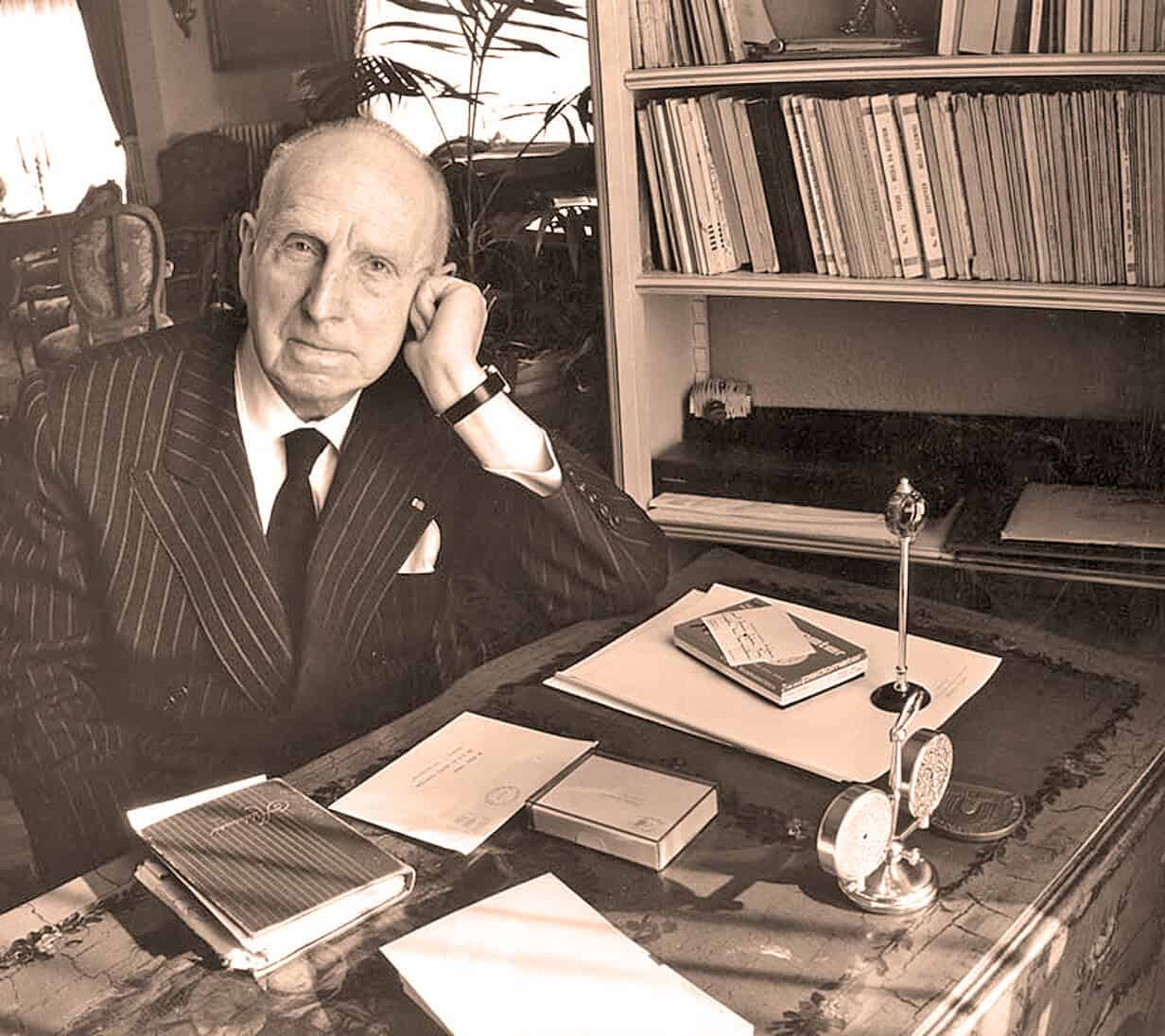 Man in vintage pinstripe suit sitting at a desk with books and notes, vintage office setting, black-and-white photo emphasizing history and archival research, related to news, history, and music archives.