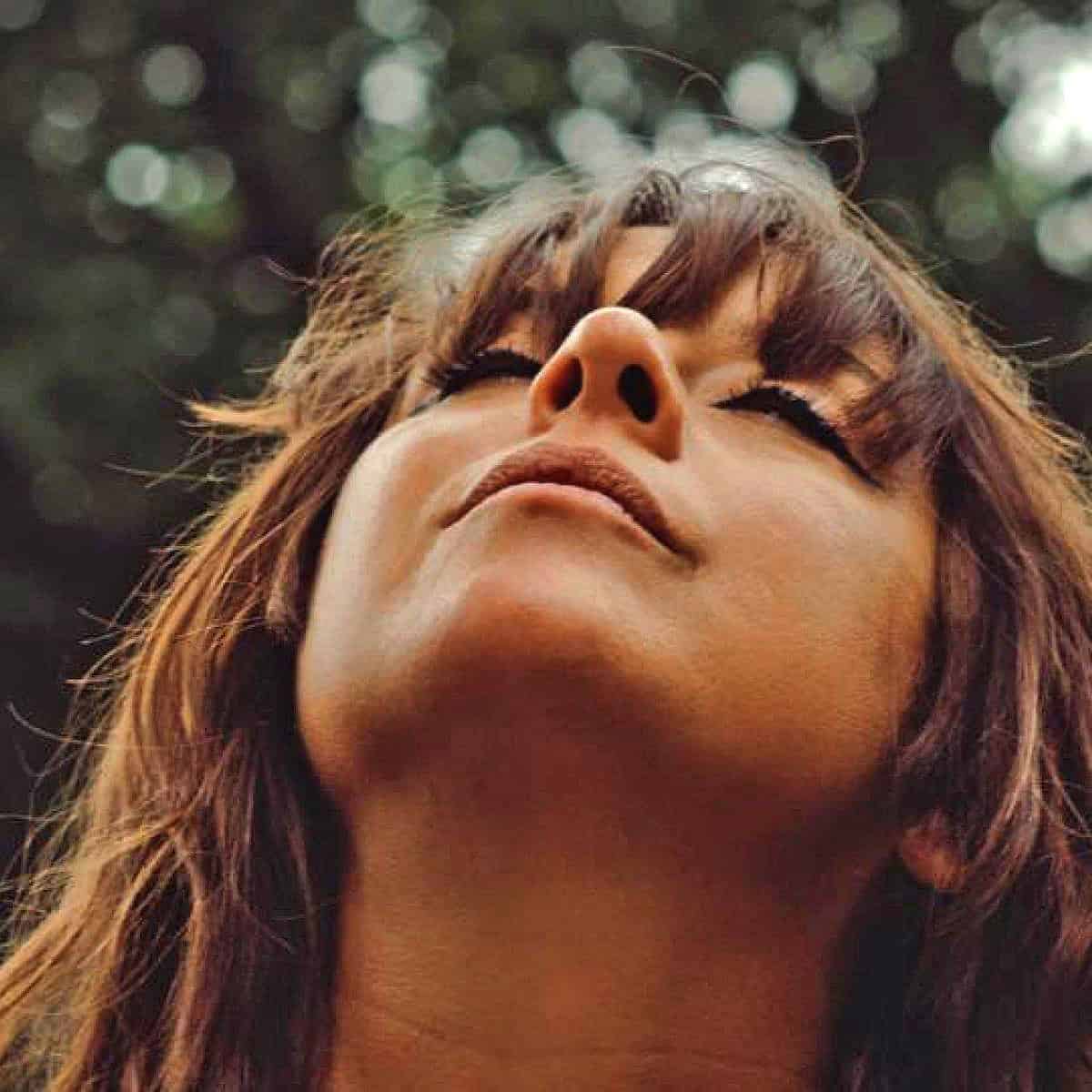 Close-up of a woman's face with her eyes closed and head tilted back, serene expression, natural outdoor setting, bokeh background, emphasizing tranquility and peacefulness for sound archive themes.