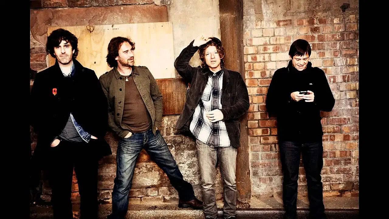 Young men in casual clothing standing against a brick wall, with one looking at a phone, capturing a candid moment in an urban or music scene.