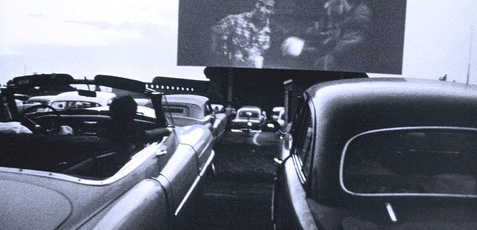 Classic vintage cars parked at drive-in theater, black and white photo, nostalgic 1950s American car culture, outdoor movie screening, historic automotive scene.