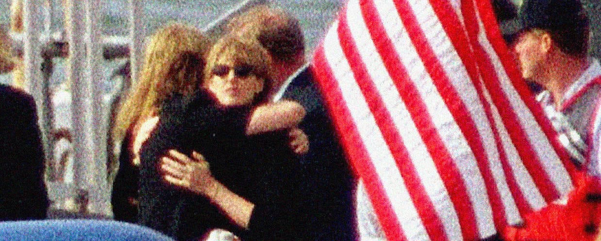 People hugging at an outdoor event under American flags, capturing a moment of reunion or celebration.