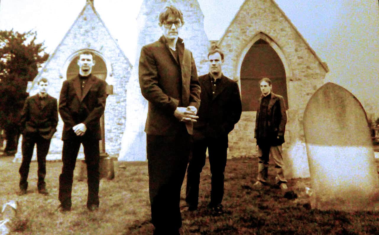 Graveyard with five men in formal attire standing near old stone church, vintage style, historical music archive, news, and sound recording context, Past Daily archive image.