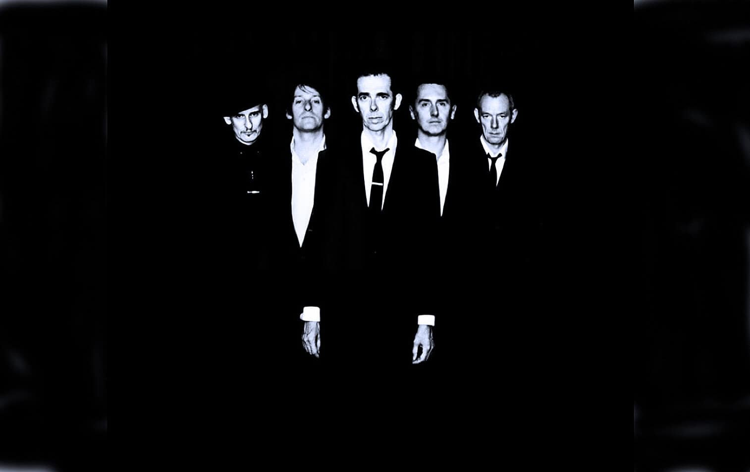 Black and white photo of a five-member band dressed in formal suits, standing against a black background, evoking classic rock or alternative music scene.
