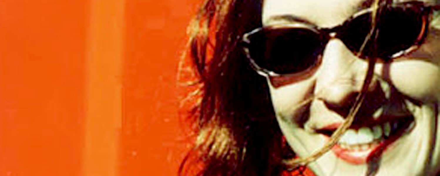Vibrant image of a woman with red hair wearing sunglasses, smiling brightly, capturing a lively and energetic atmosphere related to music, news, and history archives from Past Daily.
