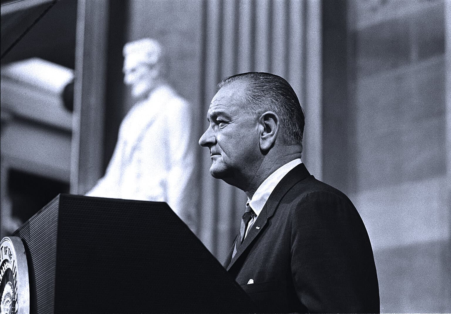 Black and white photo of a man in a suit speaking at a podium during a historic news event or political speech.