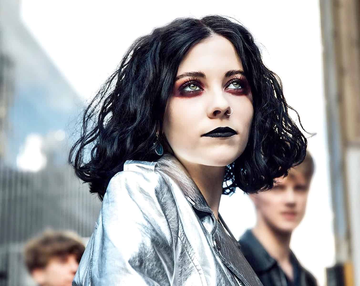 Vampire woman with black curly hair, dark makeup, silver jacket, city street background, alternative fashion, edgy style, urban environment, Gothic look, youthful attitude, music and cultural expression, nightlife scene, contemporary streetwear, music event, artistic portrait.