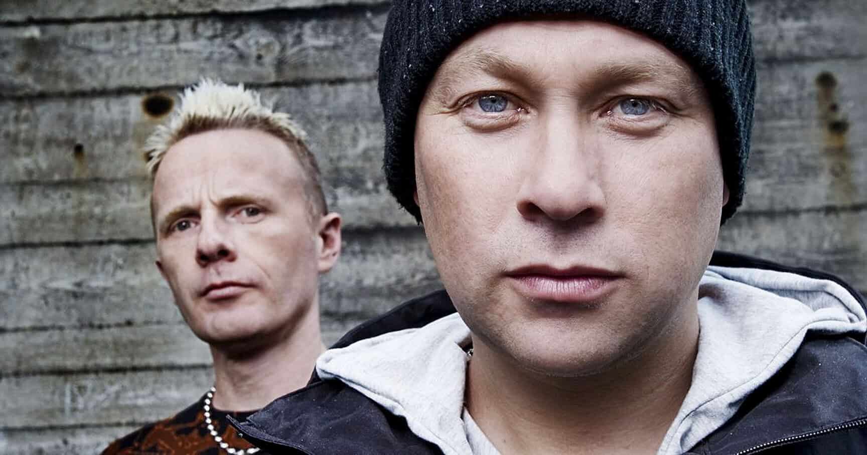 Close-up of two young men with serious expressions, standing outdoors against a weathered wooden wall, capturing street style, youth culture, and urban fashion.