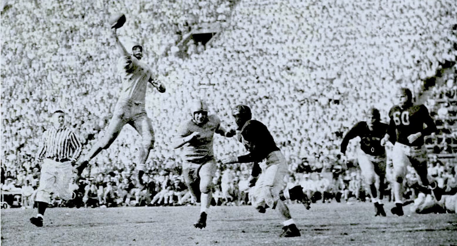 Vintage black and white football game scene with players in action during a historic game, surrounded by a large crowd in the stadium.