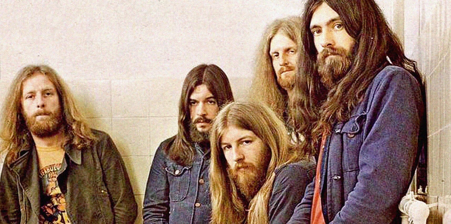 Retro rock band members with long hair and beards in casual clothing, posing in a music-related setting, representing the classic rock era.