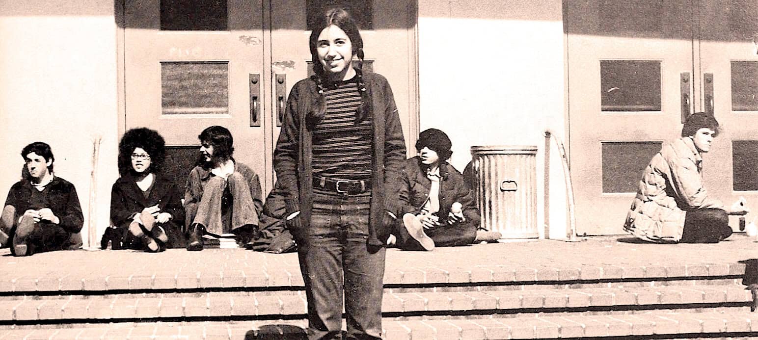 Teen girl standing on school steps in vintage black and white photo, with peers sitting behind her, capturing a moment in youth, education, and social history.