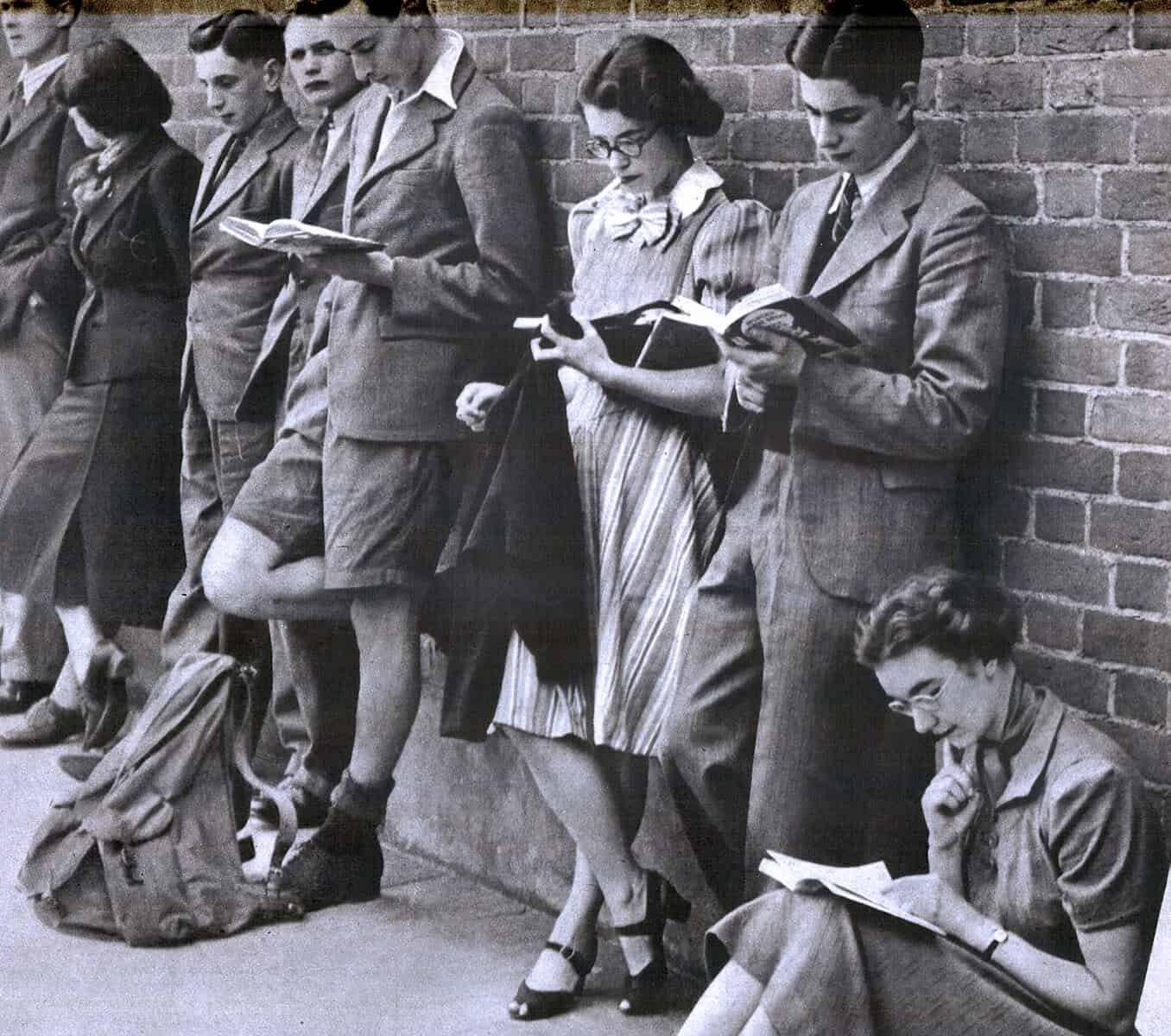 Teenagers reading books and magazines while leaning against a brick wall, showcasing youth culture and historical fashion.