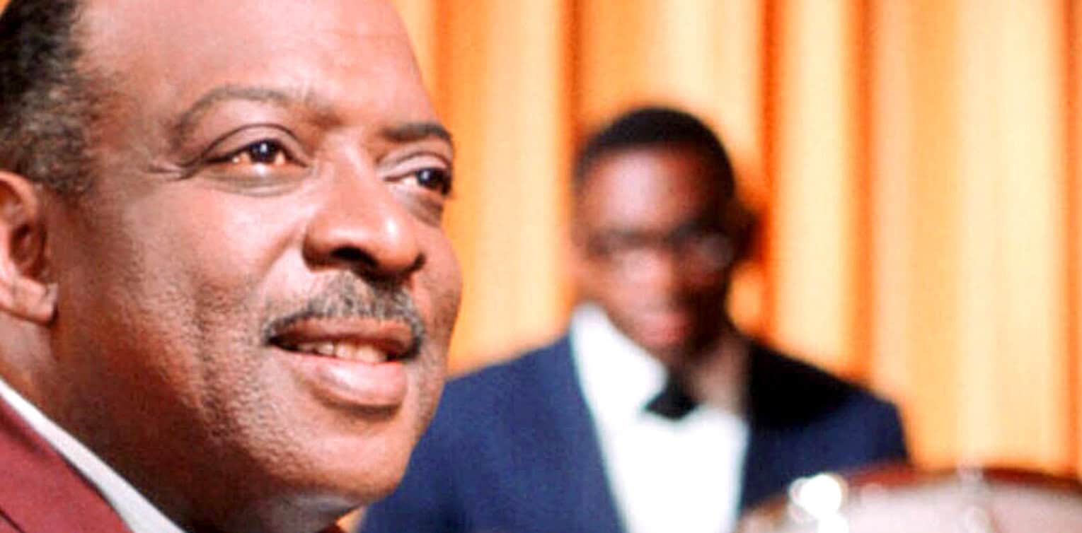 Close-up of a distinguished man with gray hair and mustache during a formal event, with another man blurred in the background, emphasizing news, history, and music archives.