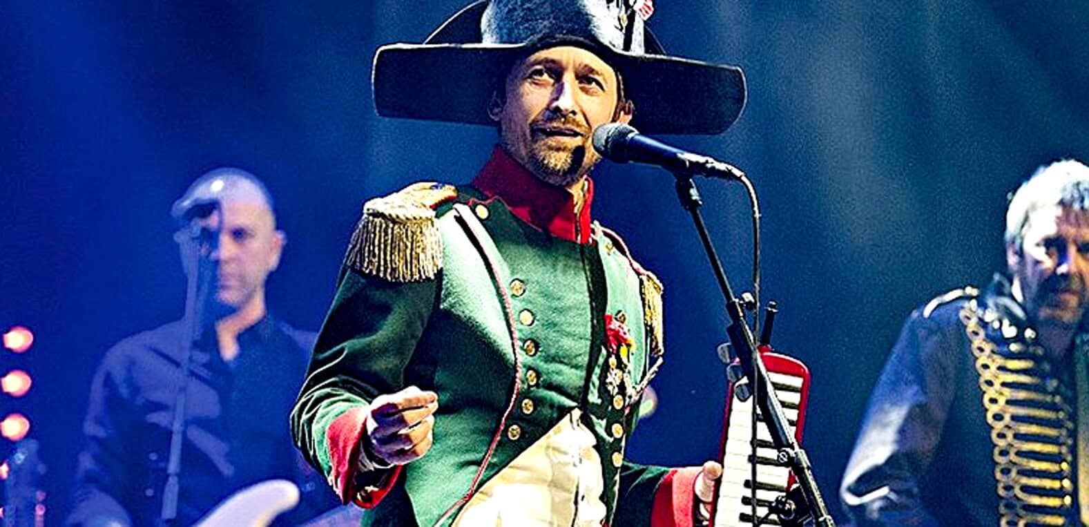 Historic musician in colorful historic costume performing on stage with microphone and band at live concert, vintage music event, musical performance, entertainment, and cultural history, 19th century style.