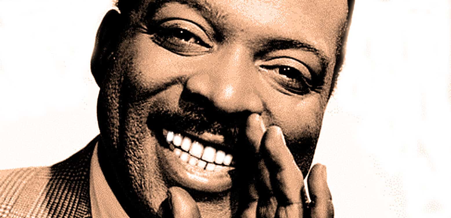 Smiling African American man with a mustache and beard, holding his hand near his mouth, showcasing a joyful expression, suitable for nostalgic music and news archives.