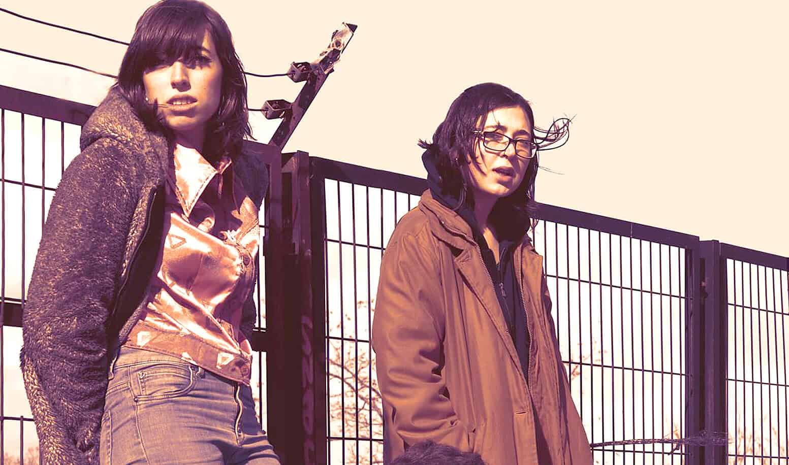 Suspenseful scene from a film with two women standing near a metal fence, capturing a moment of tension.