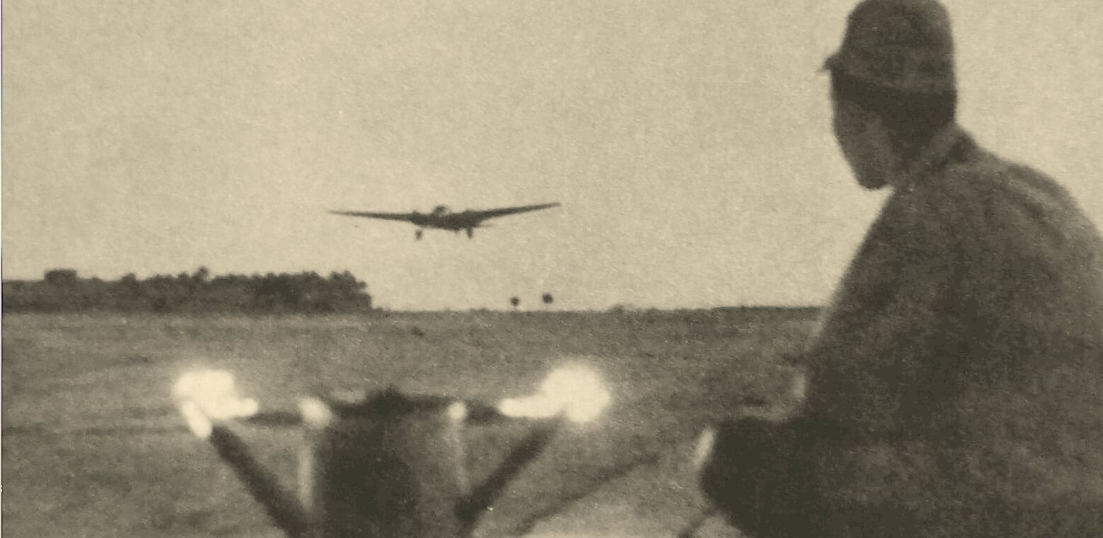 A man in military attire crouching on an open field at dusk with an aircraft flying low in the background, capturing a historic moment in aviation and military history.