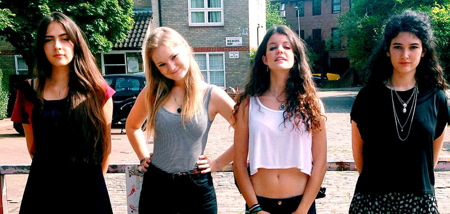 Young women standing outdoors in urban setting, casual fashion, music, youth culture, street scene, diverse expressions, lively atmosphere, vibrant summer day, trendy fashion, friendship vibe, authentic lifestyle imagery.