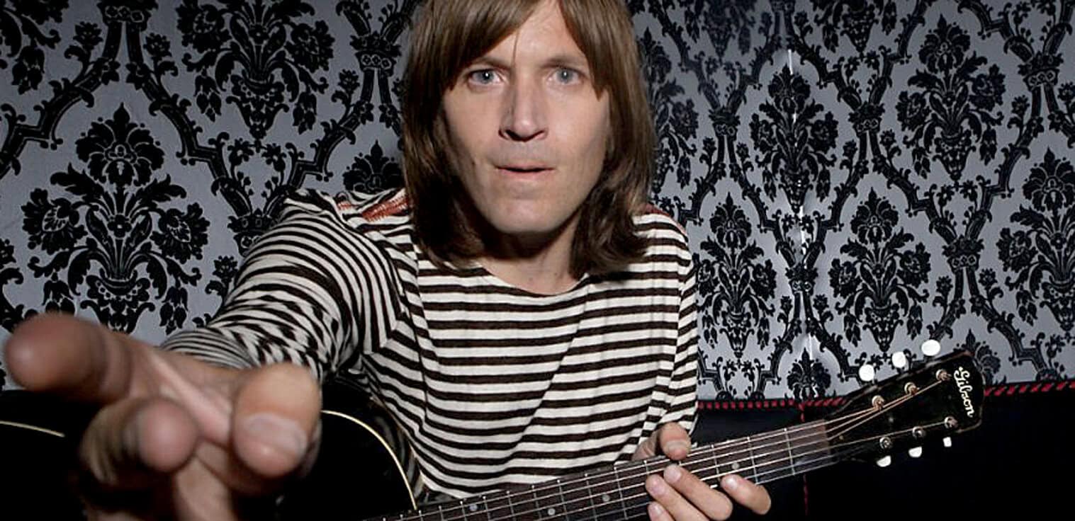 Guitarist in striped shirt reaching out towards the camera, with vintage wallpaper background, holding an acoustic guitar for music and entertainment in classic rock or acoustic performance context.