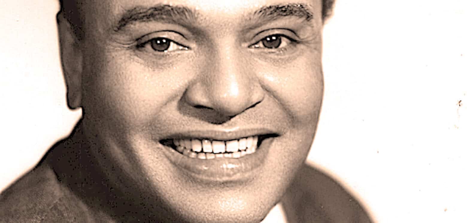 Close-up black and white photo of a smiling man with short hair, showcasing his teeth and joyful expression, representing historic news and music archive content from Past Daily.