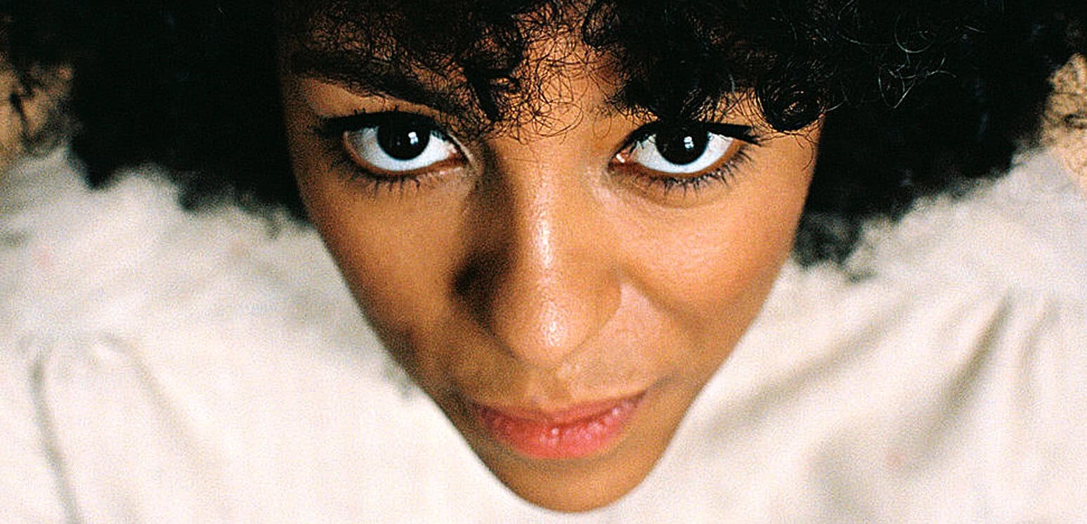 Close-up of a woman with curly hair and intense brown eyes, showcasing a confident expression with natural makeup and a soft smile, highlighting beauty and personality.