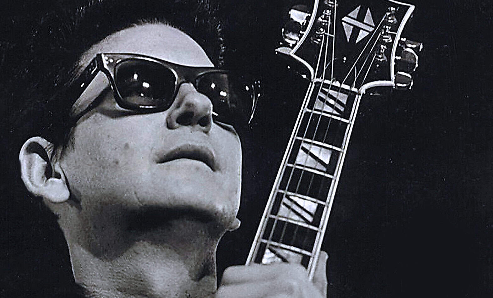 A black and white close-up of a musician wearing sunglasses and holding a guitar neck, emphasizing iconic music performance, history, and sound archives.