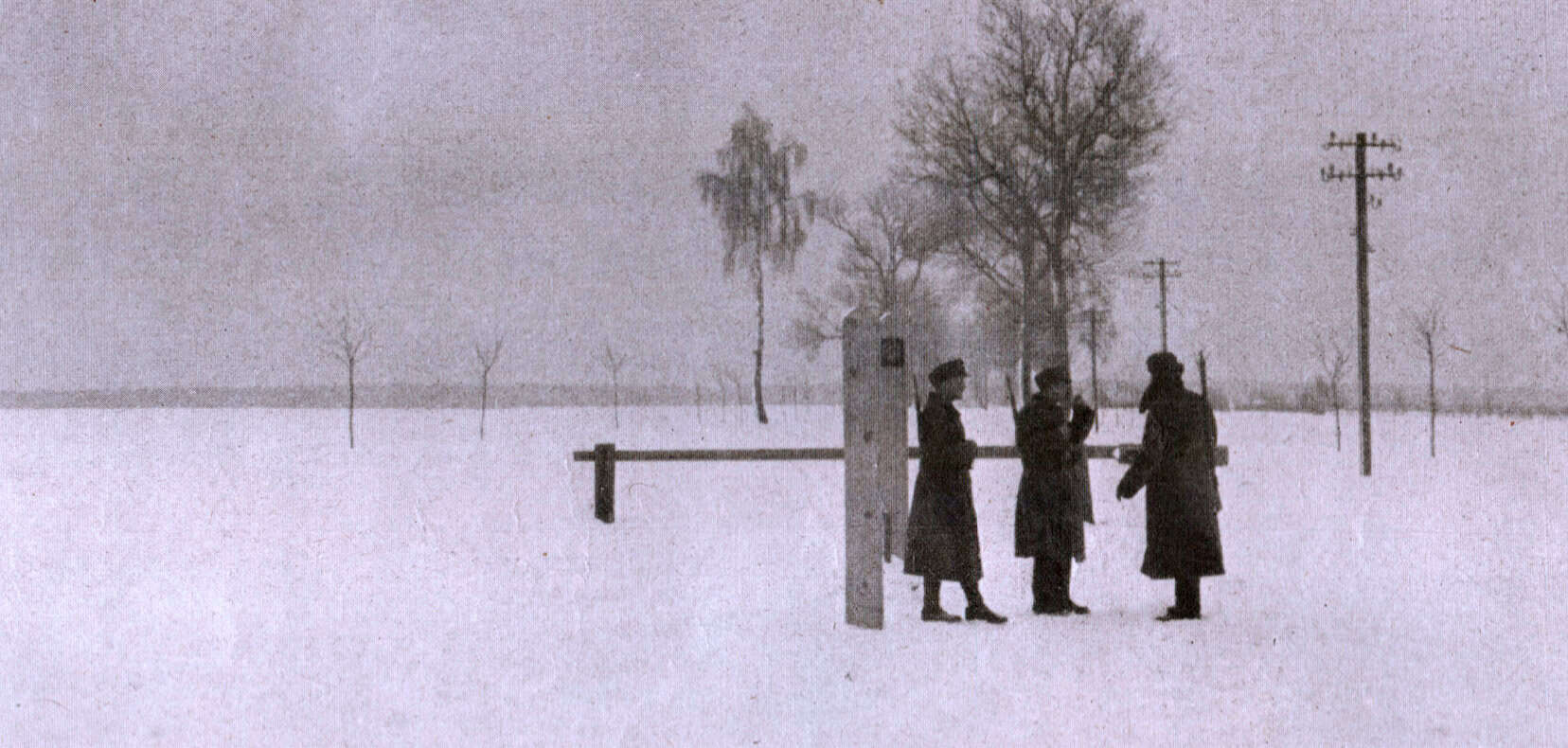 Dullsnowy landscape with three women in early 20th-century winter coats standing near a wooden gate, surrounded by barren trees and telephone poles, capturing historical winter scene.