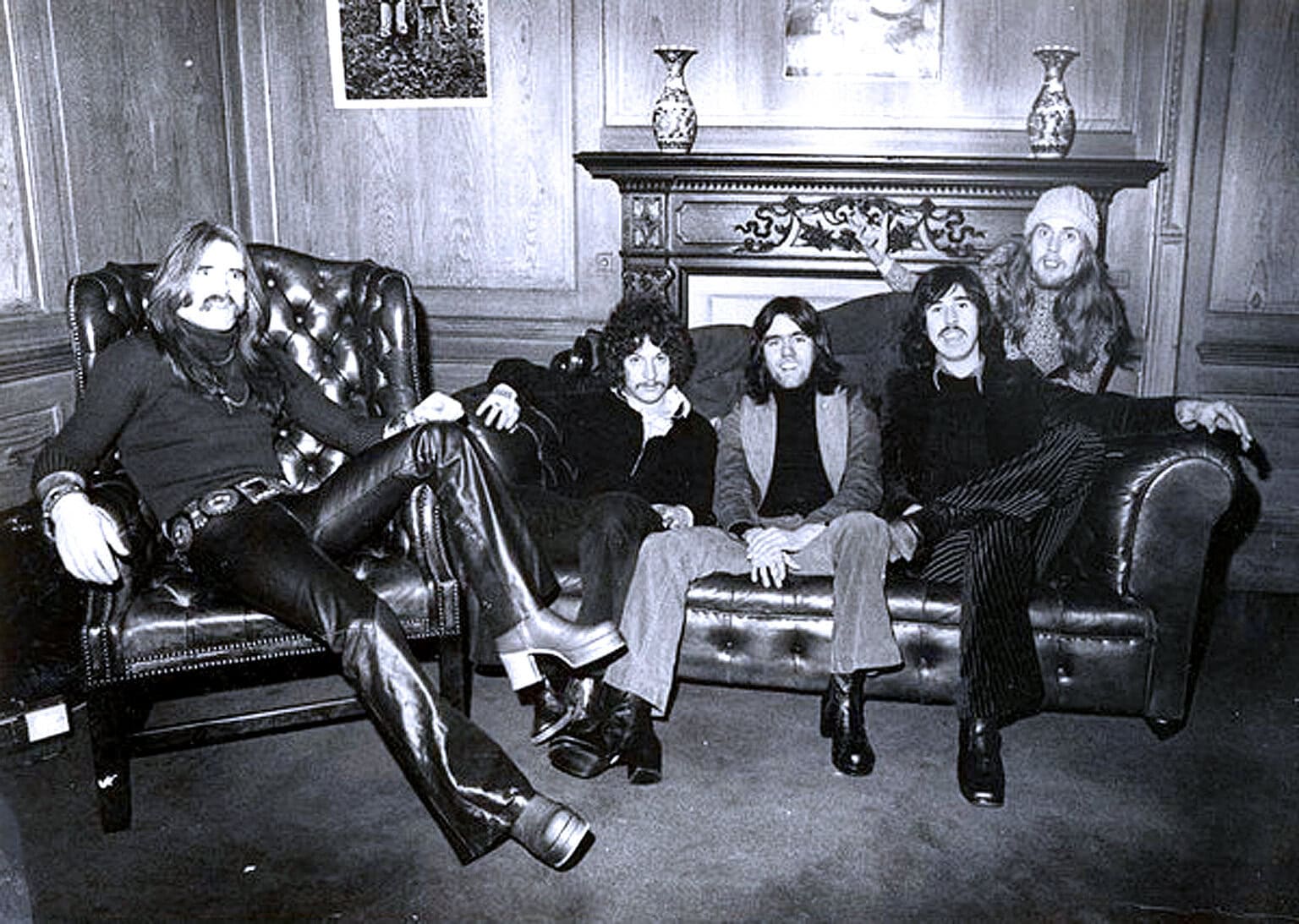 Vintage black and white photo of a classic rock band lounging on a leather sofa in a cozy, wood-paneled room, capturing the essence of 1960s or 70s music culture.
