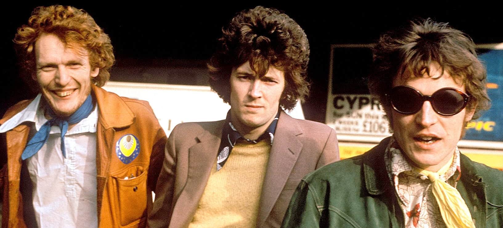 Red-haired man with a cheerful smile in vintage attire; central man with curly hair and serious expression; younger man with sunglasses and patterned shirt; retro style, iconic music and news history scene.