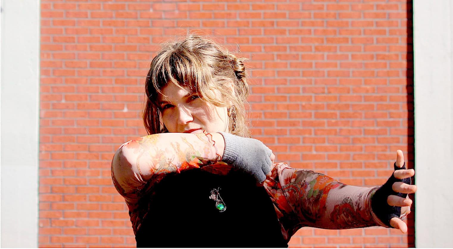 Girl with tattooed arm taking a selfie outdoors in front of a brick wall; urban street style, casual fashion, confident pose, sunlight, vibrant colors, modern accessories.