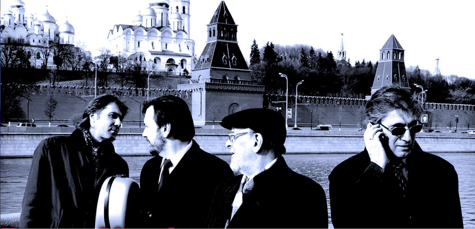 Vintage street scene with four men in conversation, historic architecture in the background, black and white photo emphasizing news, history, and music archive themes.
