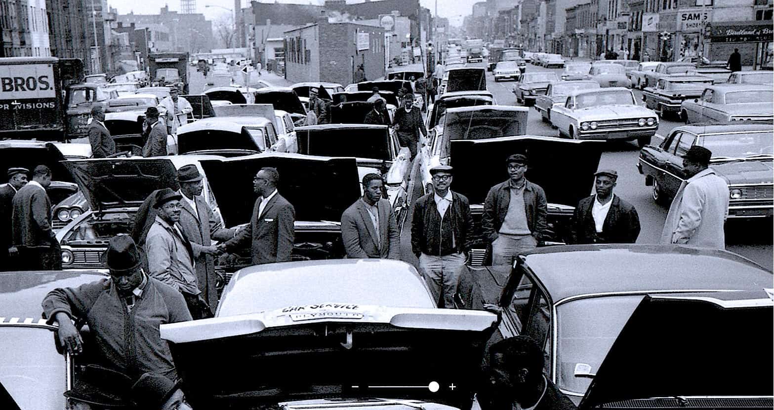 Black and white photo of a busy urban street filled with vintage cars and pedestrians during the civil rights era in the 1960s.