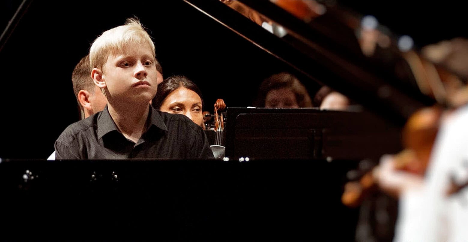 Young boy playing piano at a concert showcasing classical music, with an audience and other musicians nearby; part of a music archive highlighting live performances and musical history.