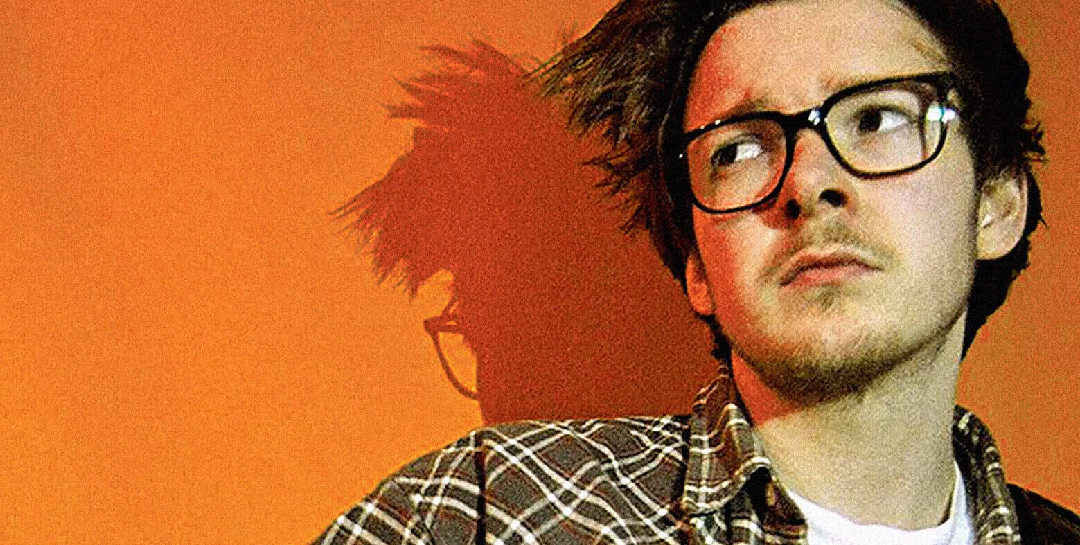 Portrait of a young man with glasses and a plaid shirt on an orange background, emphasizing music, news, history, and sound archive themes for SEO.