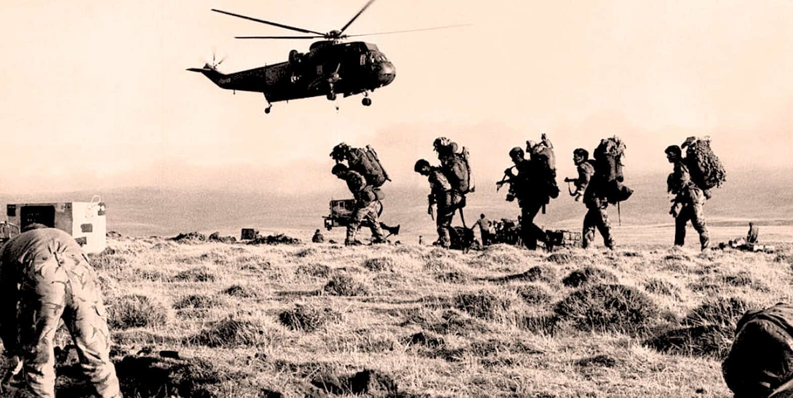 Military personnel walking across a field with a helicopter flying overhead, capturing a historic moment during wartime.