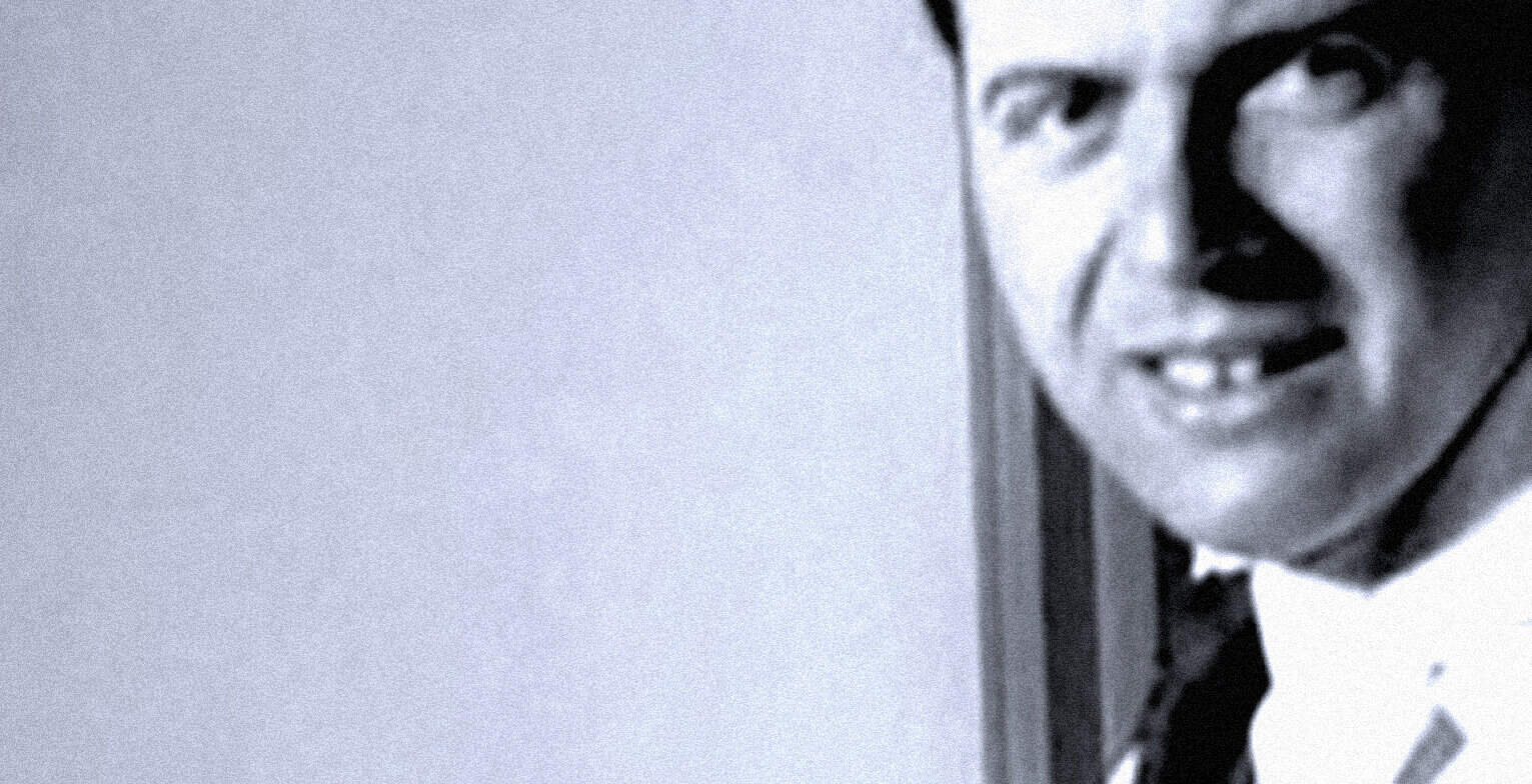 Vintage black and white photo of a smiling man peeking from behind a wall, representing historical news archives and music preservation.