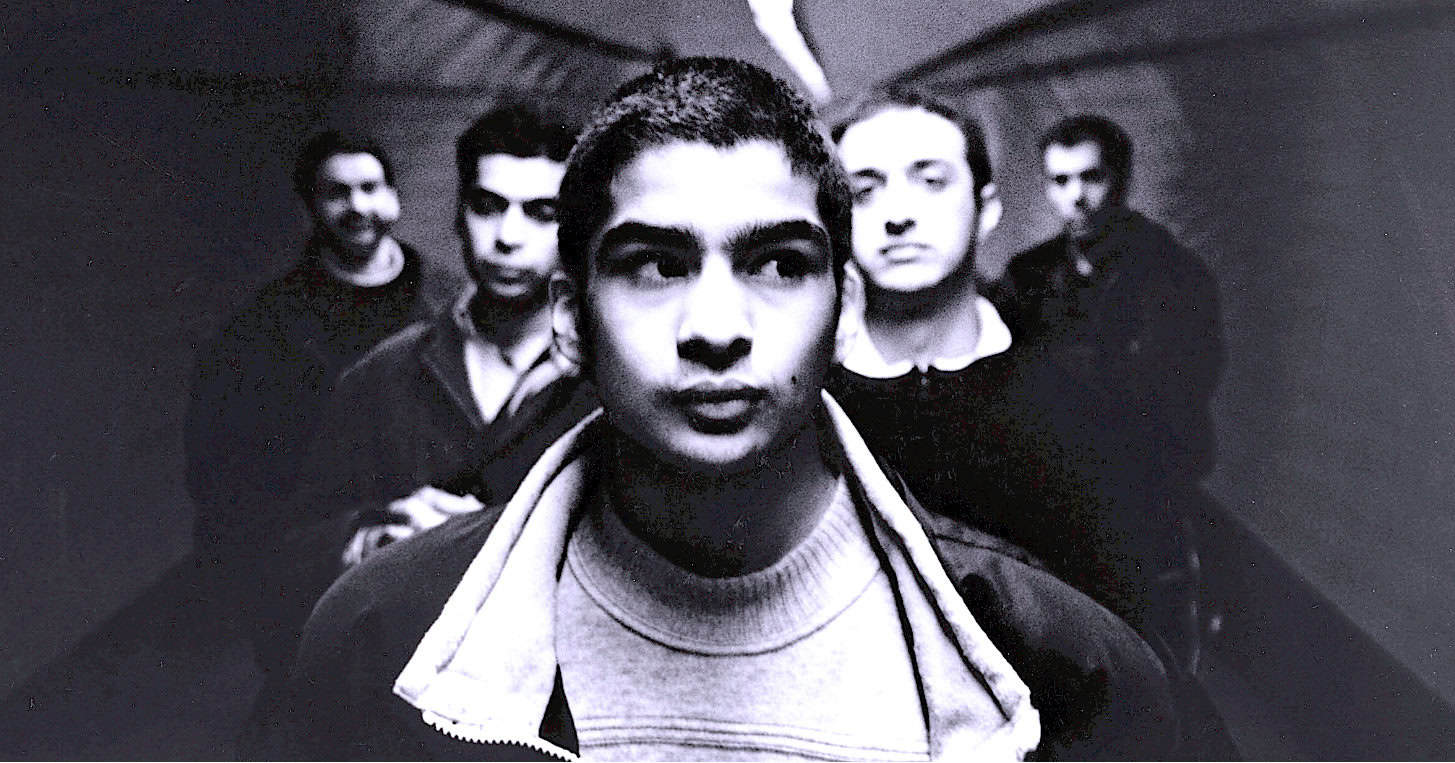Young man with a serious expression, group of men in background, black and white photo, historical image, youth culture, music archive, news archive, vintage photography, past daily, sound archive of history and music.