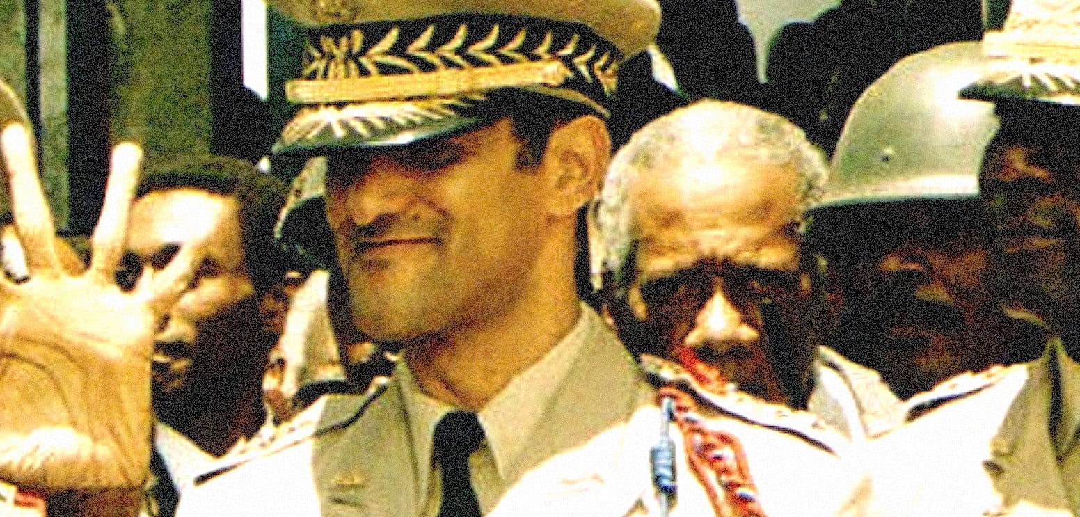 Young military officer waving during a historic event, with soldiers in helmets in the background. Image reflects news, history, and military moments captured in the sound archive.