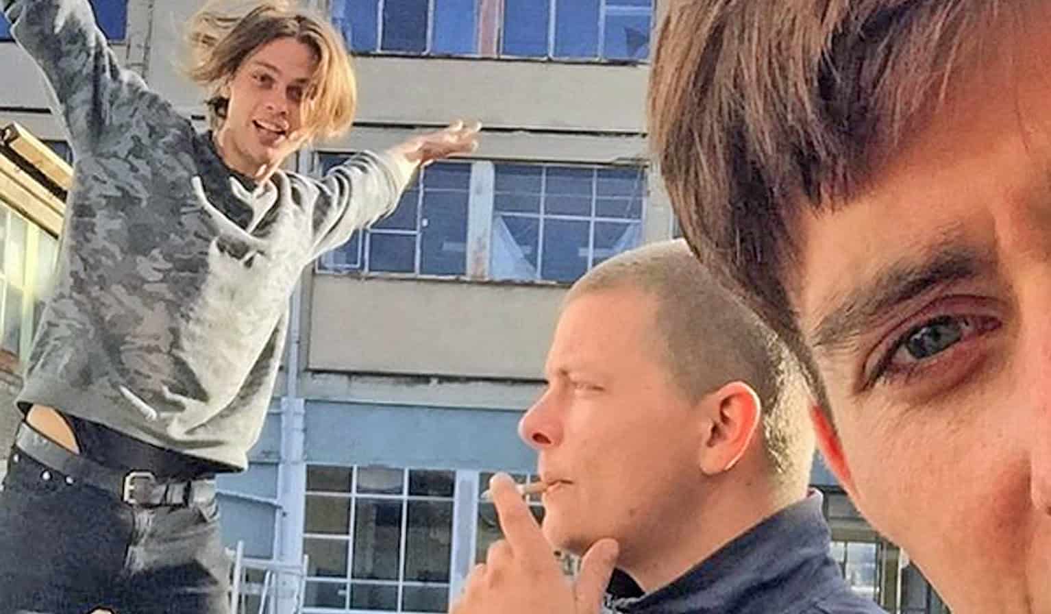 Unexpected outdoor candid of three young men hanging out outdoor urban setting, casually dressed, lively atmosphere, friendship, modern lifestyle, urban culture, social gathering, youthful energy, candid photography, community life, band or friends gathering, candid shot, street photography, urban environment, casual fashion, social interaction, authentic moments.