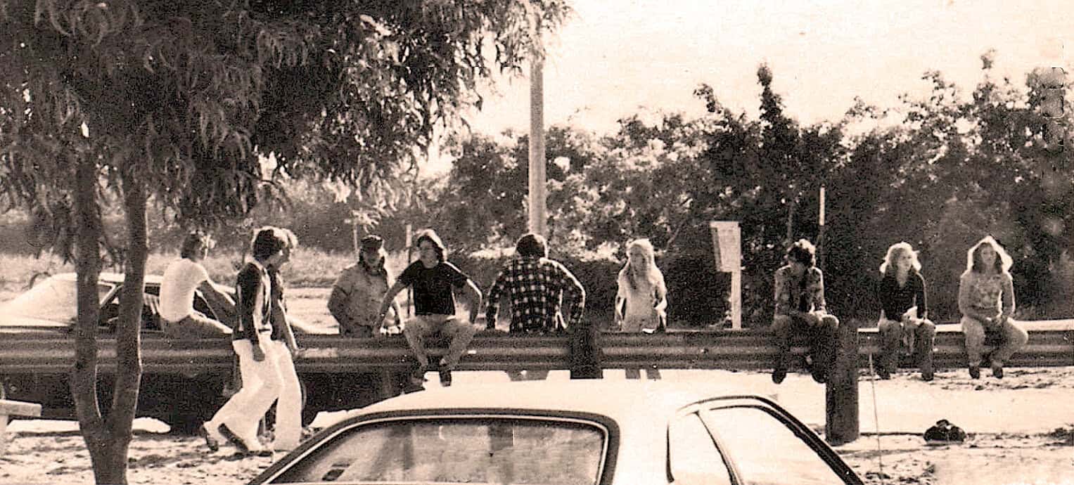 People sitting on a guardrail near trees and a vintage car in a sepia-toned photo.