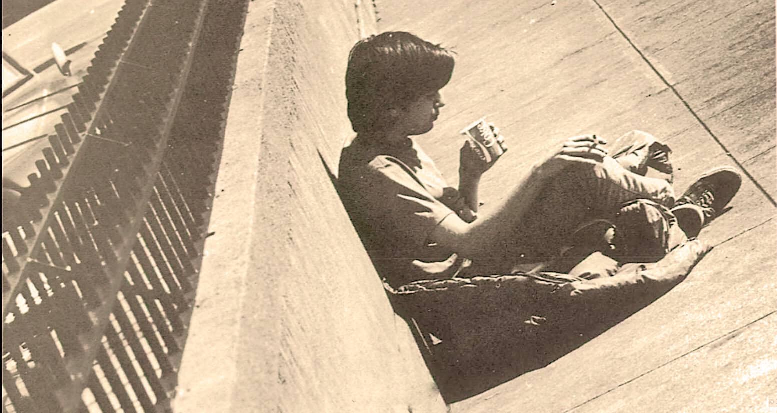 A young boy sitting on the sidewalk next to a fence, holding a can and drinking from it, in a nostalgic black-and-white photo representing street life and childhood moments.