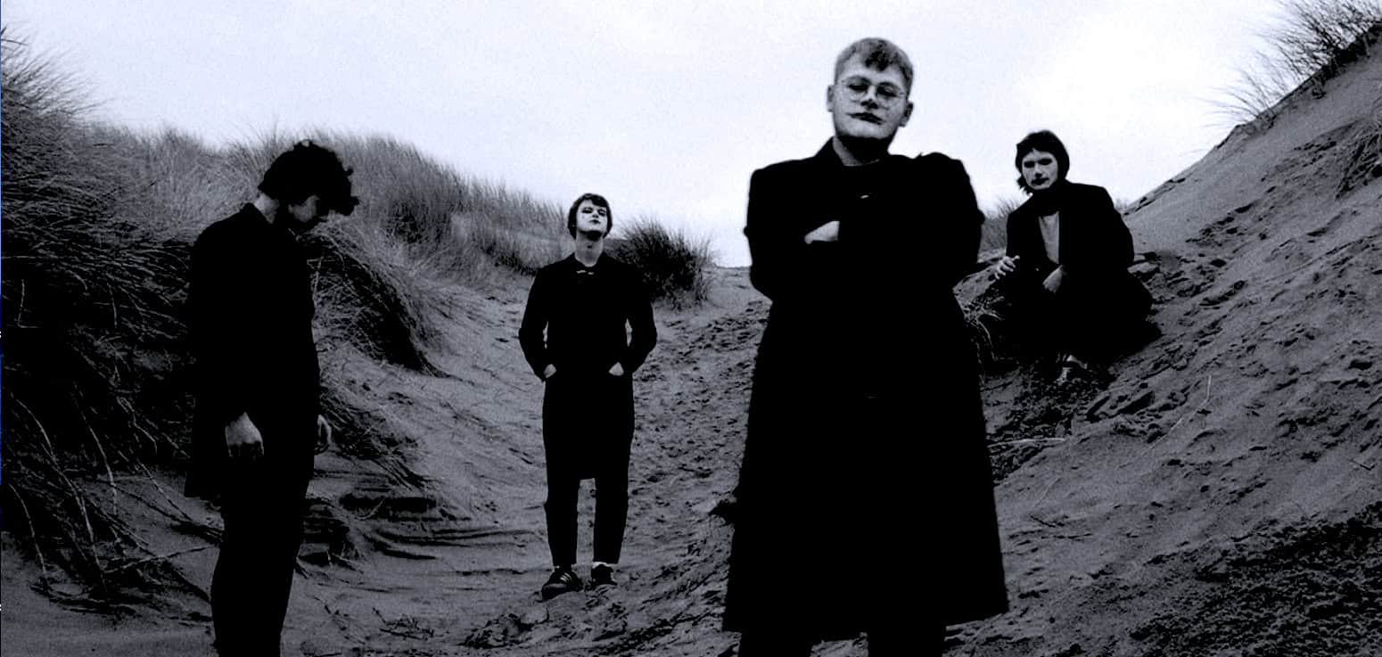 Black and white photo of four young men in stylish dark clothing on a sandy dune, showcasing alternative and indie fashion, used for vintage style music and cultural archive.